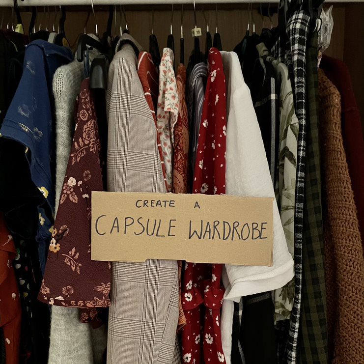 Image shows a wardrobe rail with a mixture of clothes. A cardboard sign along the shirt sleeves reads: Create a Capsule Wardrobe.