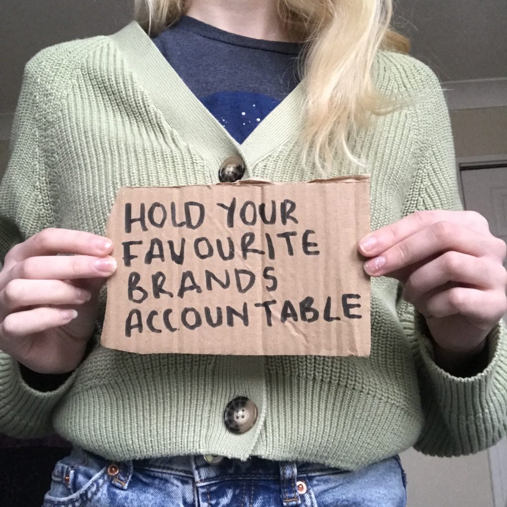 Image of a person in a green cardigan, holding a cardboard sign with "Hold your favourite brands accountable' written on it