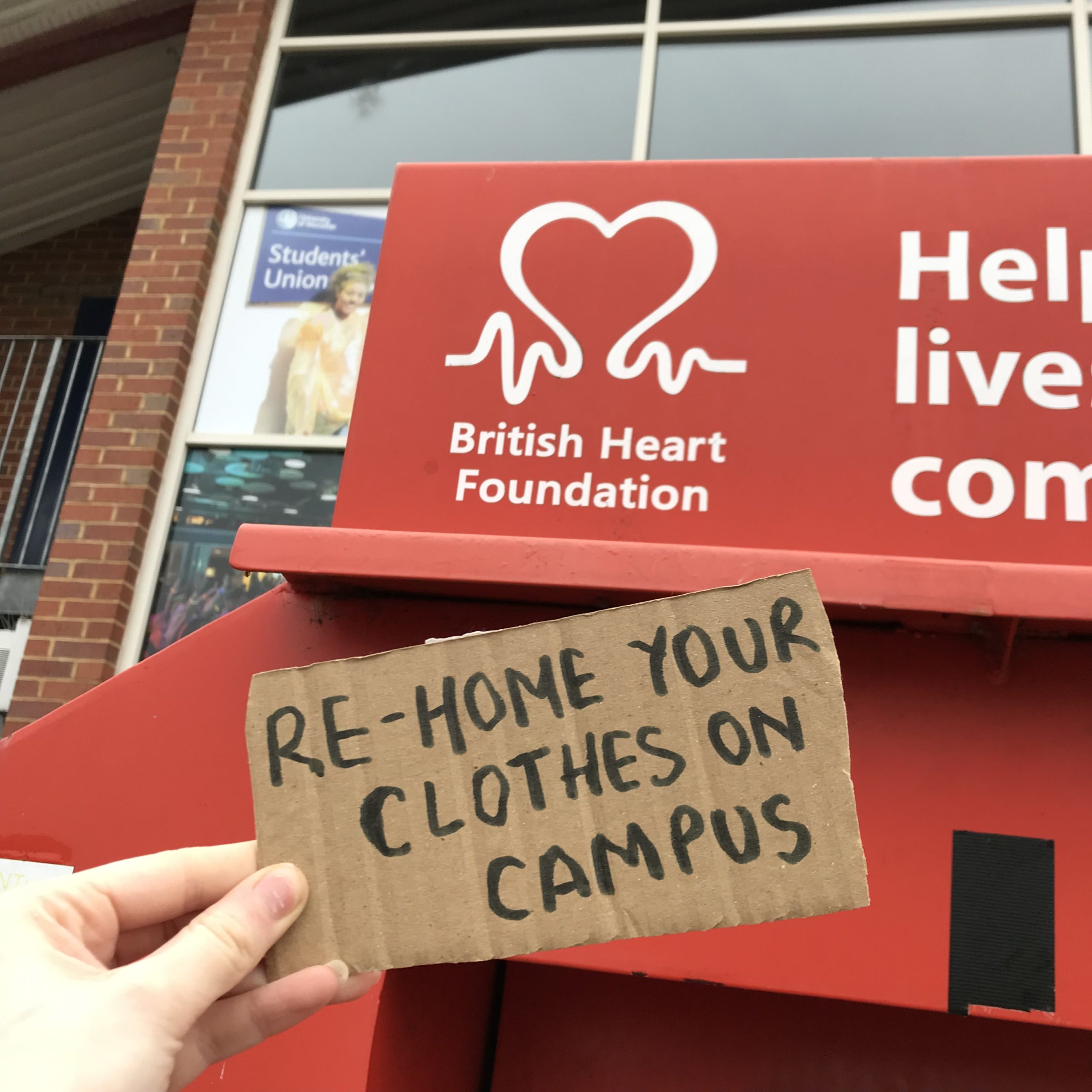 Image in the foreground of a piece of cardboard, with 'Re-home your clothes on campus' written on it. In the background is a British Heart Foundation donation bank, located outside the student union