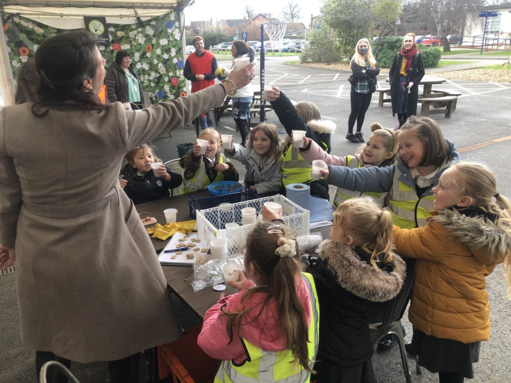 Children from Oldbury Primary School standing around a wooden table, holding up white paper cups bird feed like they are giving a toasting, a woman in a long brown coat stands at the head of the table and holds her white cup to toast them back.