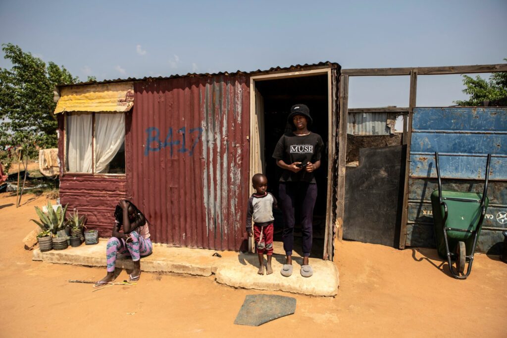 Two people stood and one person sat next to a corrugated iron structure