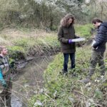 Three students, one in waders standing in Duck Brook, a second holding a tray with water samples in, the thrird recording data on a phone